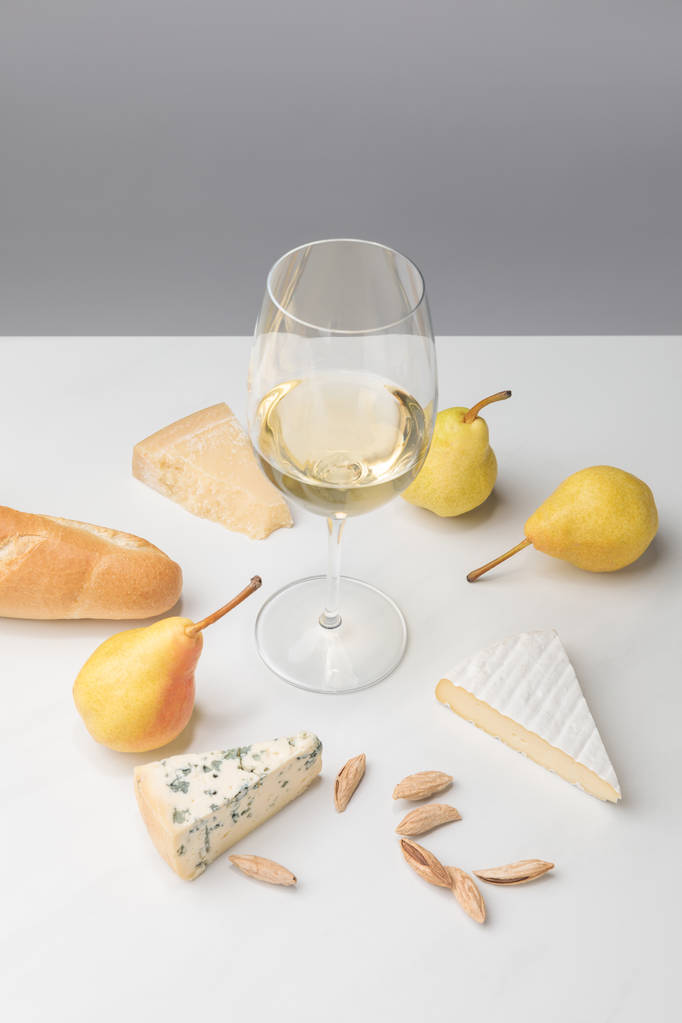 Korkeat näkymät viinilasiin, jota ympäröivät päärynät, manteli, patonki ja erilaiset juustot harmaalla
 - Valokuva, kuva