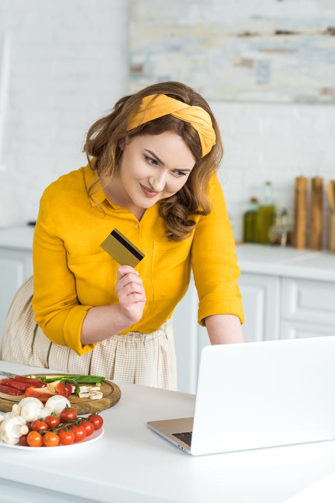 smiling beautiful woman holding credit card and looking at laptop at kitchen - Photo, Image