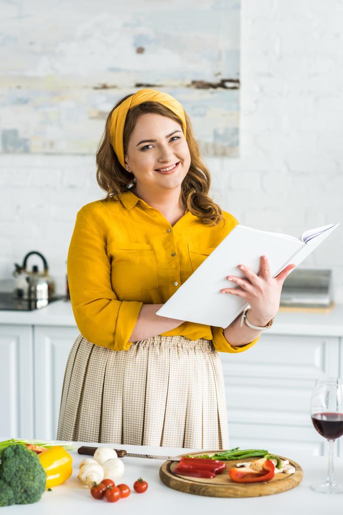 sorrindo bela mulher segurando livro de receitas e olhando para a câmera na cozinha
 - Foto, Imagem