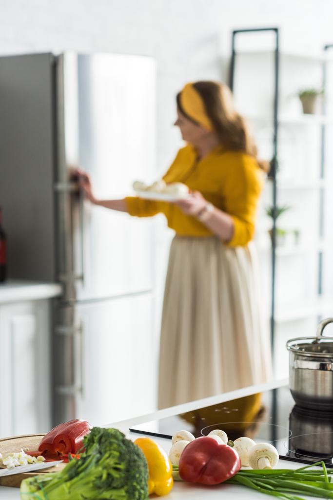 женщина берет тарелку из холодильника с овощами на переднем плане на кухне
 - Фото, изображение