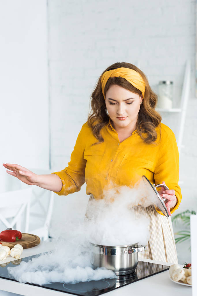 красивая женщина держит крышку и глядя на пар от кастрюли на электрической плите на кухне
 - Фото, изображение