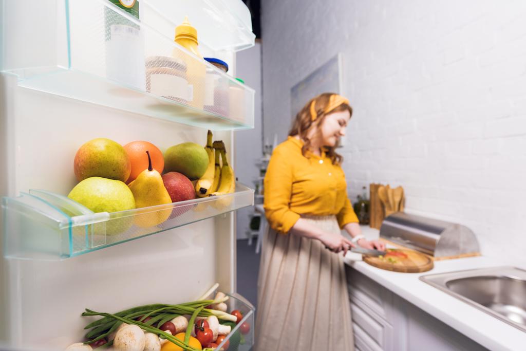 schöne Frau schneidet Gemüse in der Küche mit Kühlschrank im Vordergrund - Foto, Bild