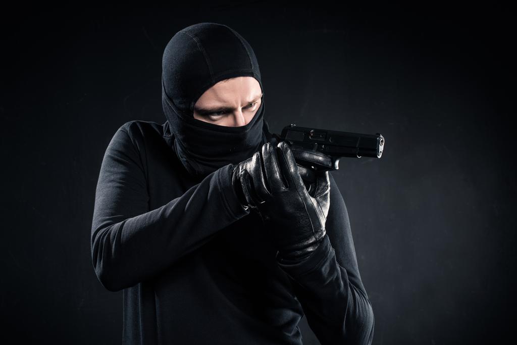 Burglar in balaclava aiming with gun on black - Photo, Image