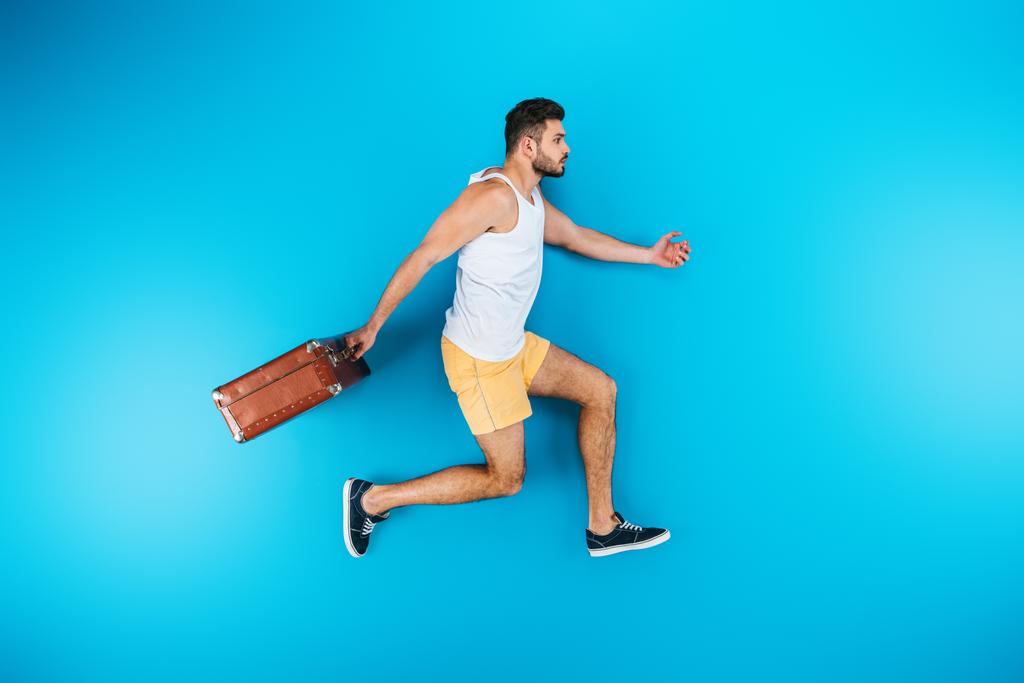 vista completa de la longitud del joven guapo sosteniendo la maleta y corriendo en azul
 - Foto, imagen