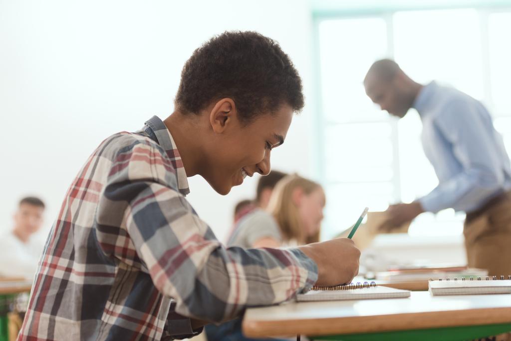 Seitenansicht eines lächelnden afrikanisch-amerikanischen Jungen, der im Lehrbuch mit Lehrer und Klassenkameraden auf der Rückseite schreibt  - Foto, Bild