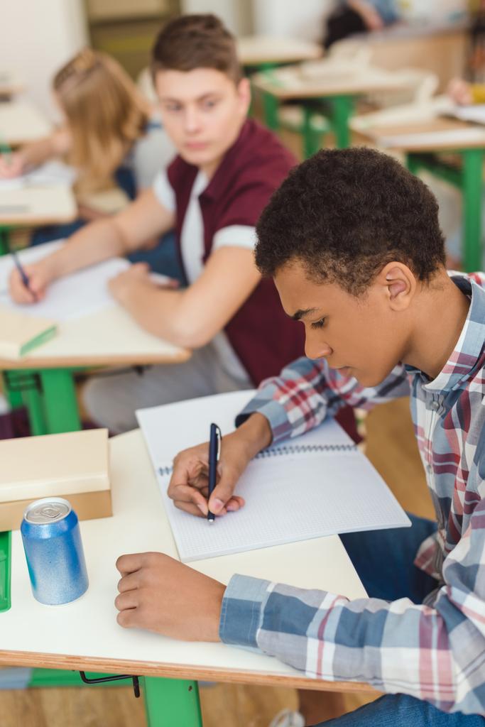 Vue élevée de l'écriture afro-américaine adolescent écolier dans le manuel et camarades de classe sur derrière
  - Photo, image