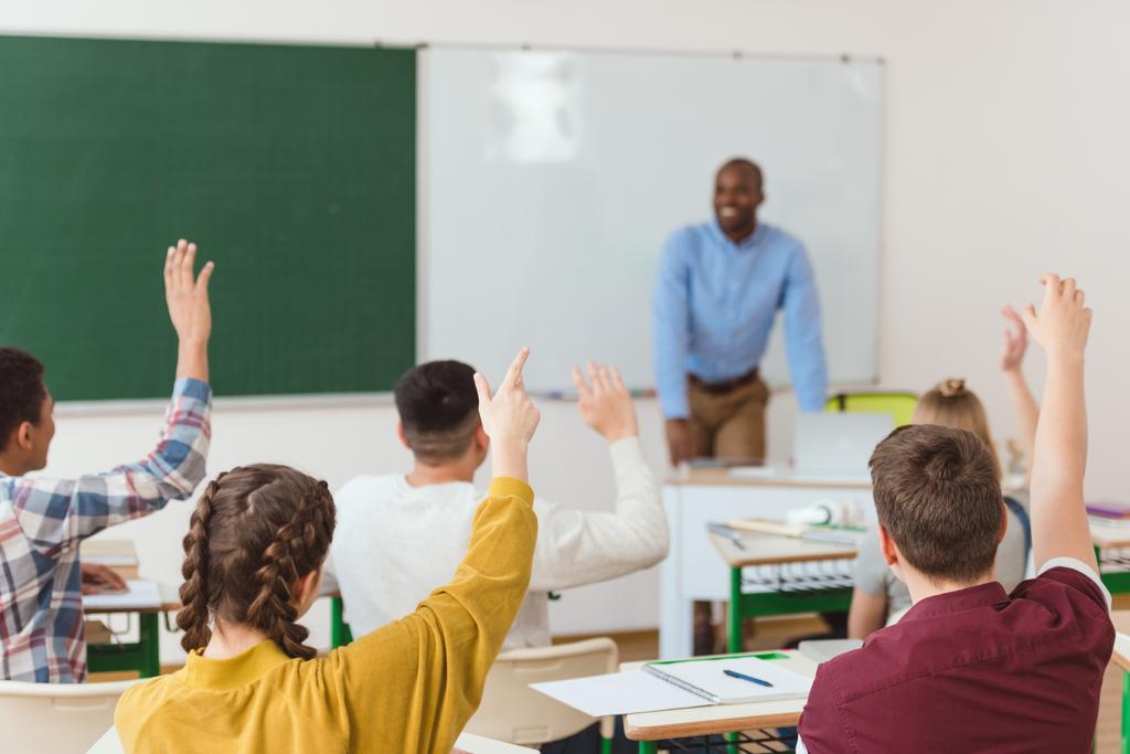Vista trasera de los estudiantes de secundaria con los brazos arriba y el profesor afroamericano en el aula
  - Foto, Imagen