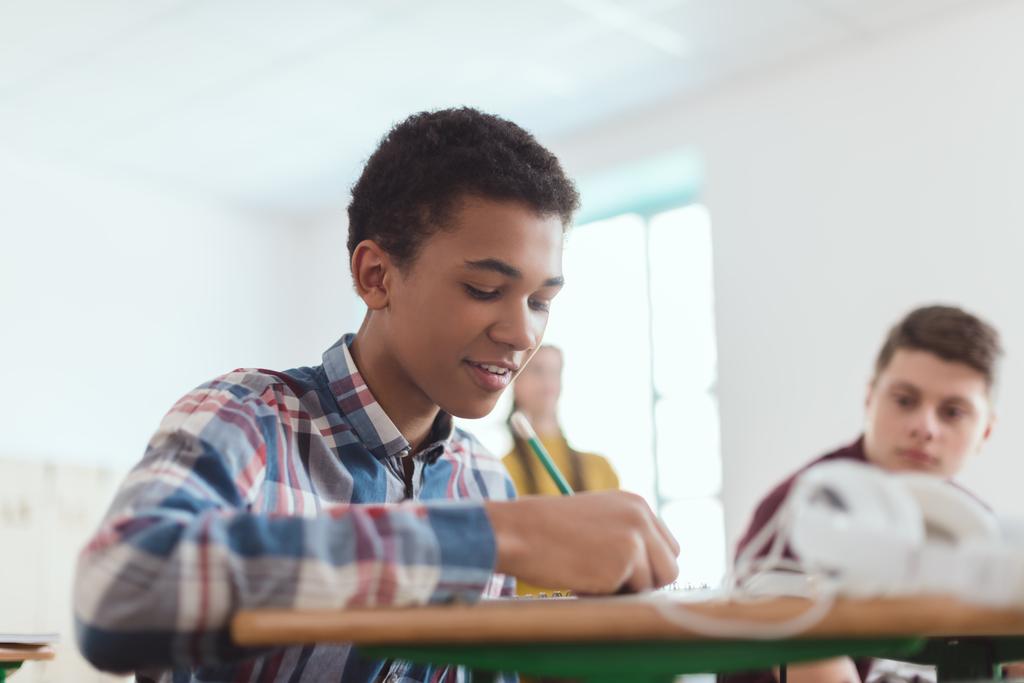 Низкий угол обзора африканского подростка старшеклассник пишет в учебнике и одноклассники позади
  - Фото, изображение