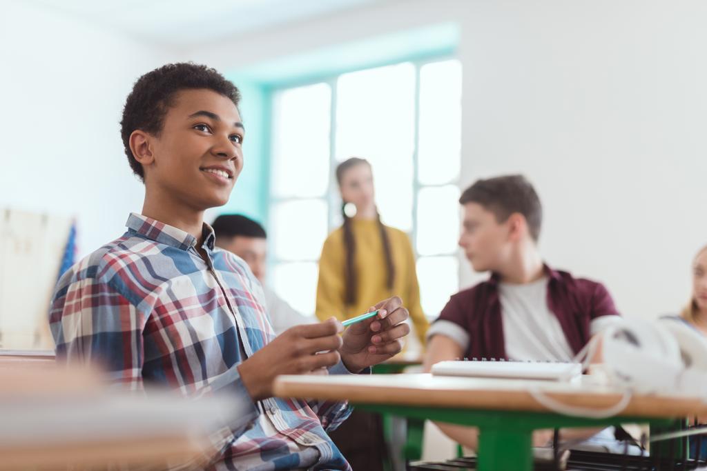 Vista basso angolo di africano americano studente di scuola superiore che tiene la matita in mano e compagni di classe seduti dietro
 - Foto, immagini