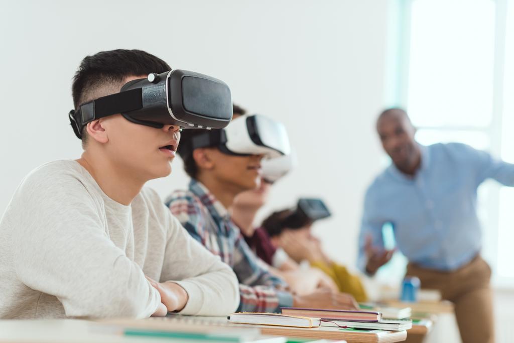 Vista lateral de escolares multiculturales que usan auriculares de realidad virtual y un profesor conversador parado detrás
 - Foto, Imagen