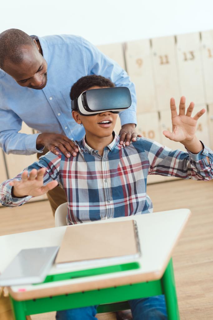 仮想現実のヘッドセットを使用して少年の肩を保持しているアフリカ系アメリカ人の教師  - 写真・画像