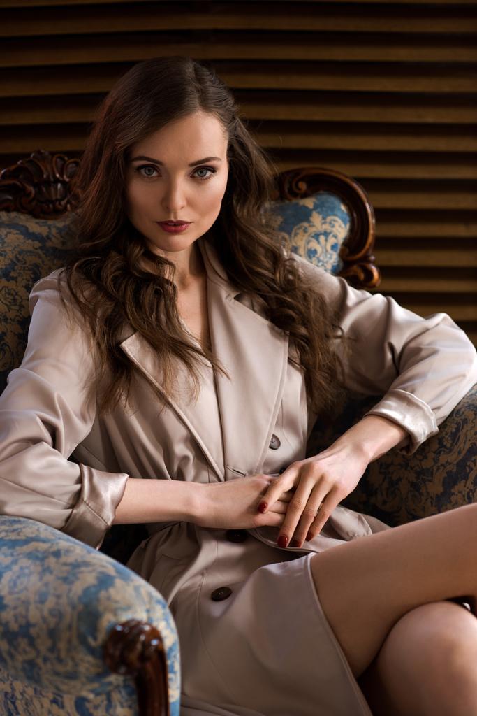 肘掛け椅子に座ってエレガントなトレンチ コートの魅力的なブルネットの女性 - 写真・画像