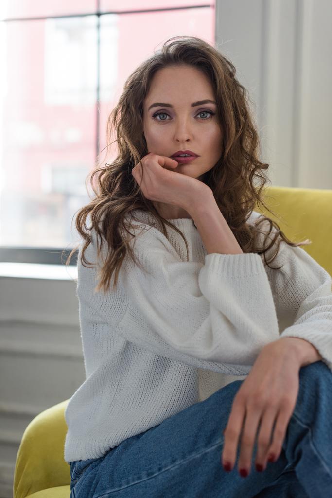 portrait of tender brunette girl in white sweater and jeans sitting on armchair and looking at camera  - Photo, Image