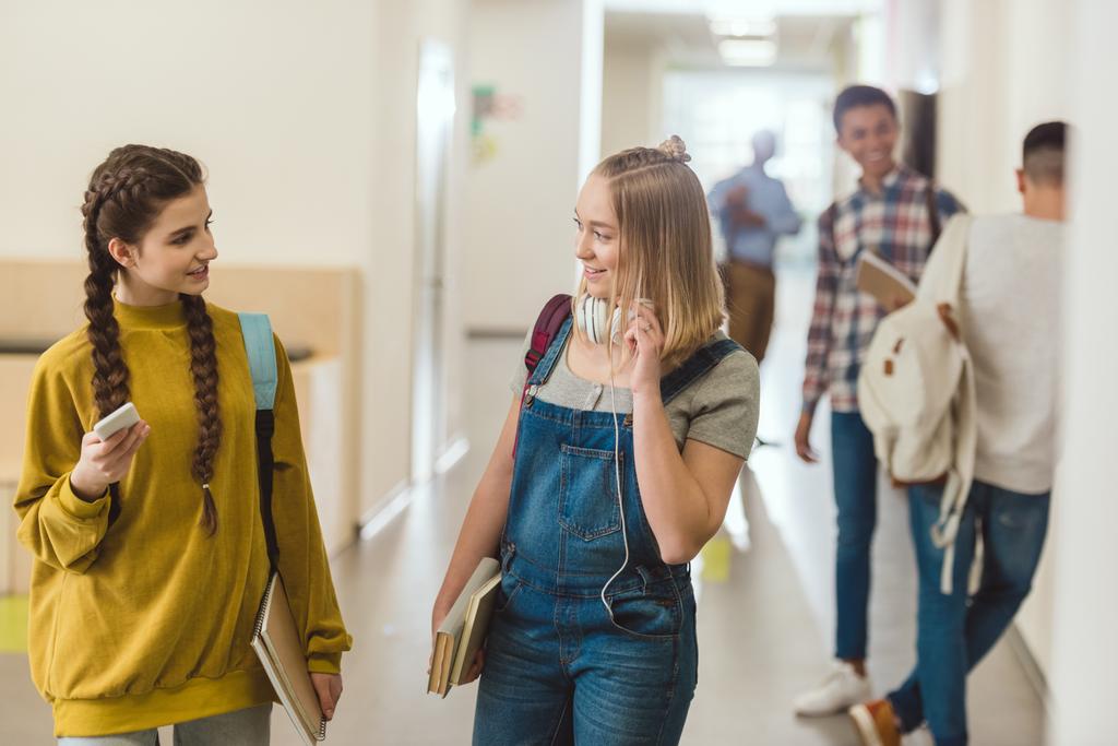 Teini-ikäiset koulutytöt kävelemässä koulun käytävällä yhdessä ja juttelemassa
 - Valokuva, kuva