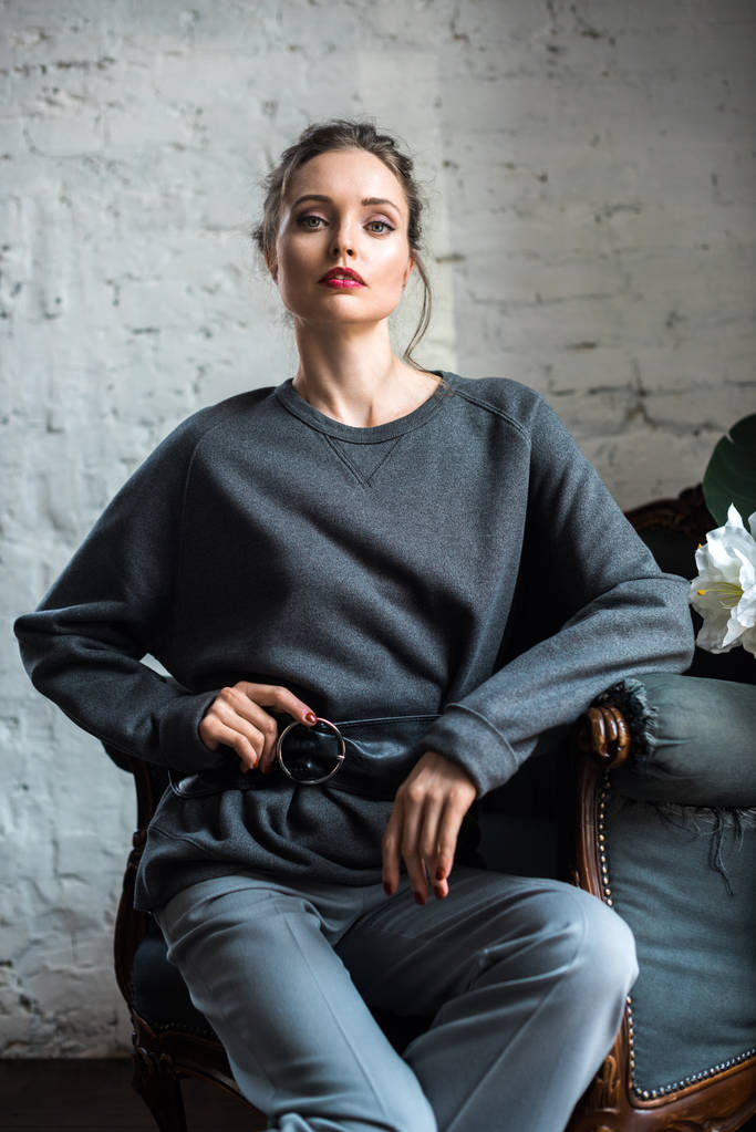 portrait of beautiful young woman in fashionable grey clothes sitting on armchair and looking at camera - Photo, Image