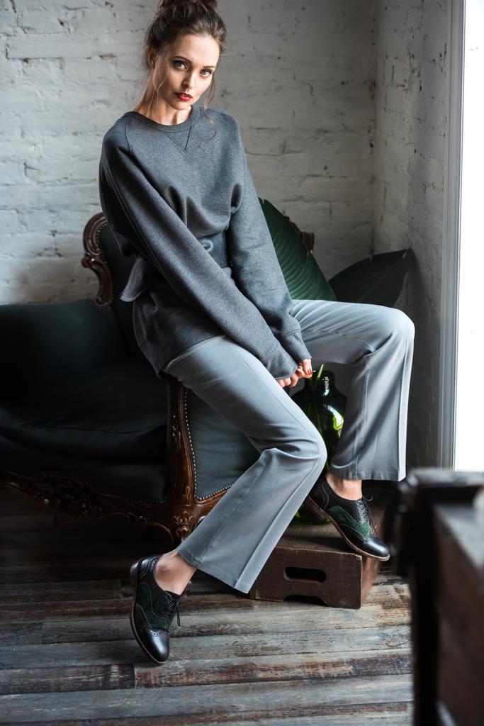 mooie brunette meisje in trendy outfit zittend op een stoel en camera kijken - Foto, afbeelding