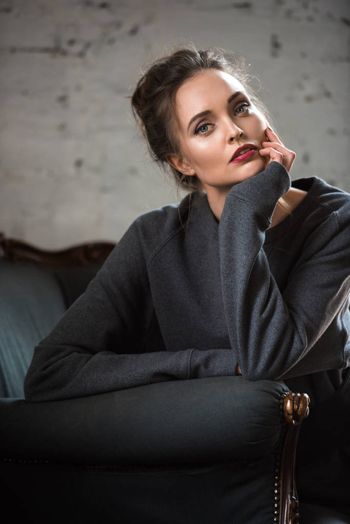 attraktive brünette Frau im trendigen Pullover sitzt auf einem Sessel und blickt in die Kamera  - Foto, Bild