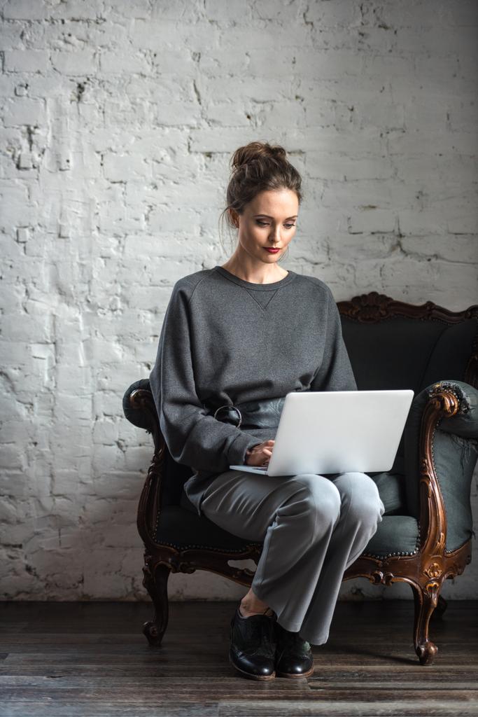 bela mulher morena em roupa elegante usando laptop enquanto sentado na poltrona
 - Foto, Imagem