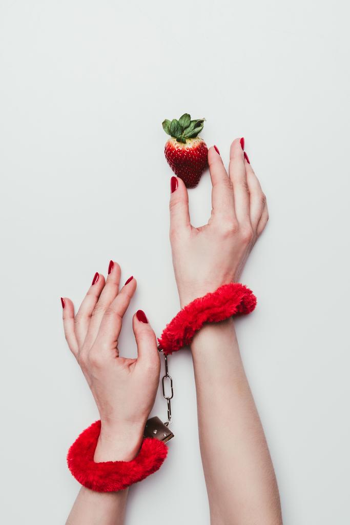 Женские руки в красных пушистых наручниках тянутся за клубникой, изолированной на белом
 - Фото, изображение