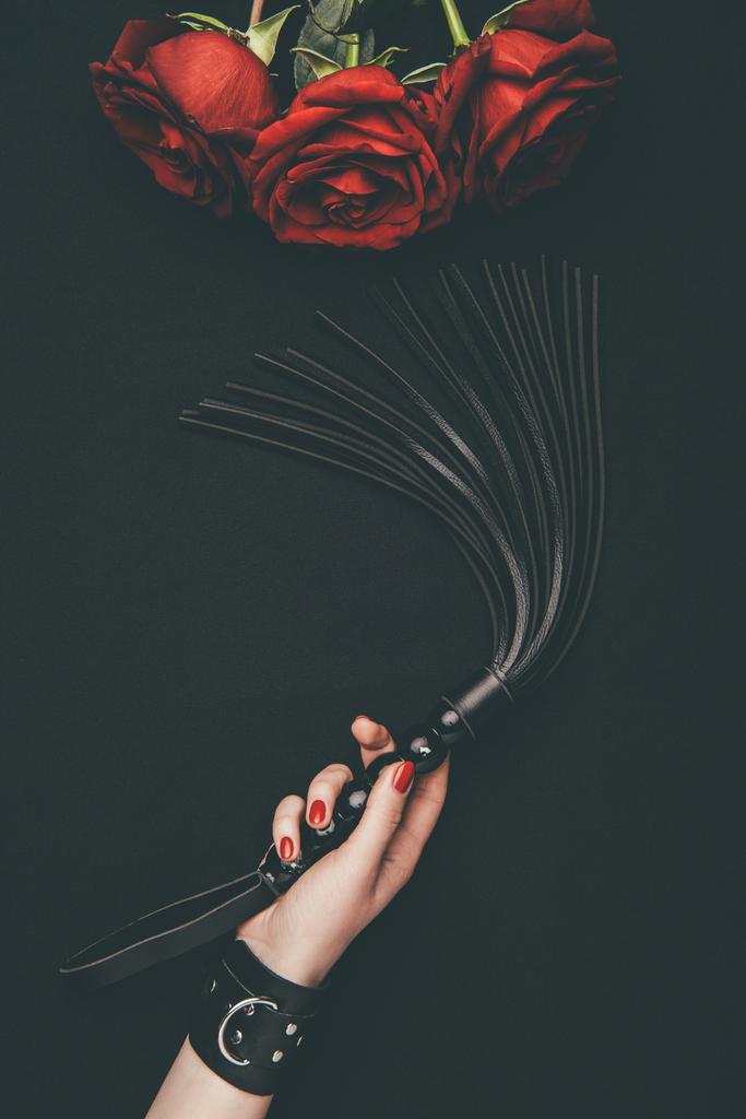 Látigo de azotes de cuero negro en mano femenina por rosas rojas aisladas en negro
 - Foto, imagen