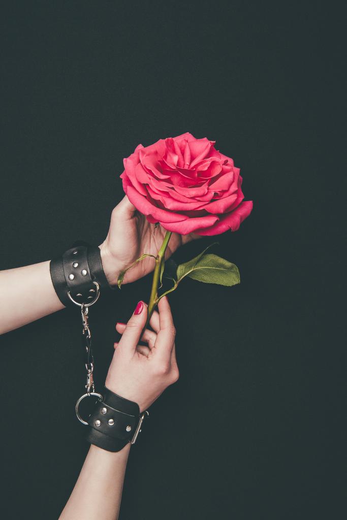 Frauenhände in Lederhandschellen, die Rosenblüten isoliert auf Schwarz halten - Foto, Bild