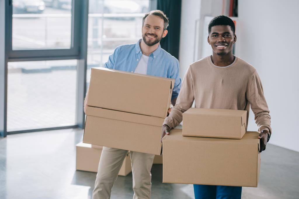 счастливые мультиэтнические мужчины, держащие картонные коробки и улыбающиеся в камеру во время переезда
 - Фото, изображение