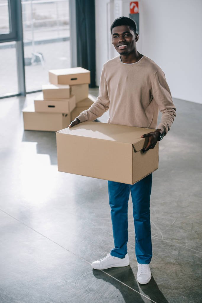young african american man holding cardboard box and smiling at camera during relocation - Photo, Image