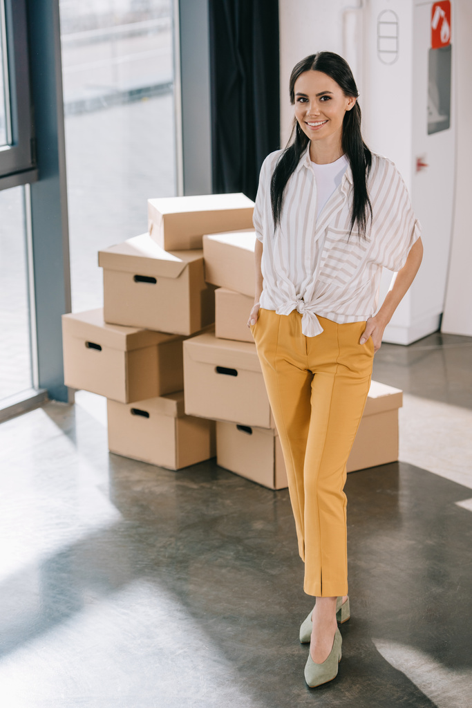 Gelukkig jonge zakenvrouw permanent met handen in de zakken en lachend op camera tijdens het verhuizen in nieuw kantoor  - Foto, afbeelding
