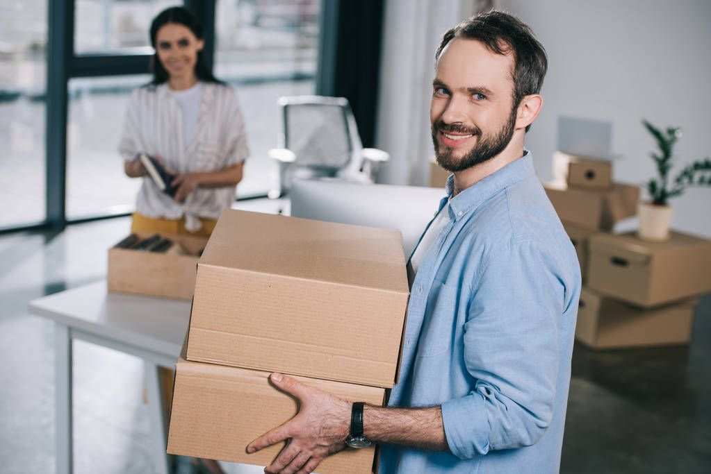 pohledný vousatý muž držící kartonových krabic a usmívá se na kameru při přemístění s kolegyně v nové kanceláři - Fotografie, Obrázek