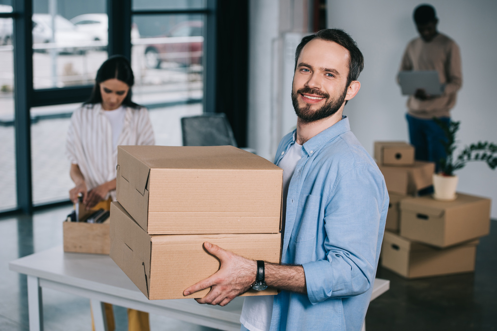 man holding cardboard boxes and smiling at camera while relocating with colleagues at new workplace  - Photo, Image