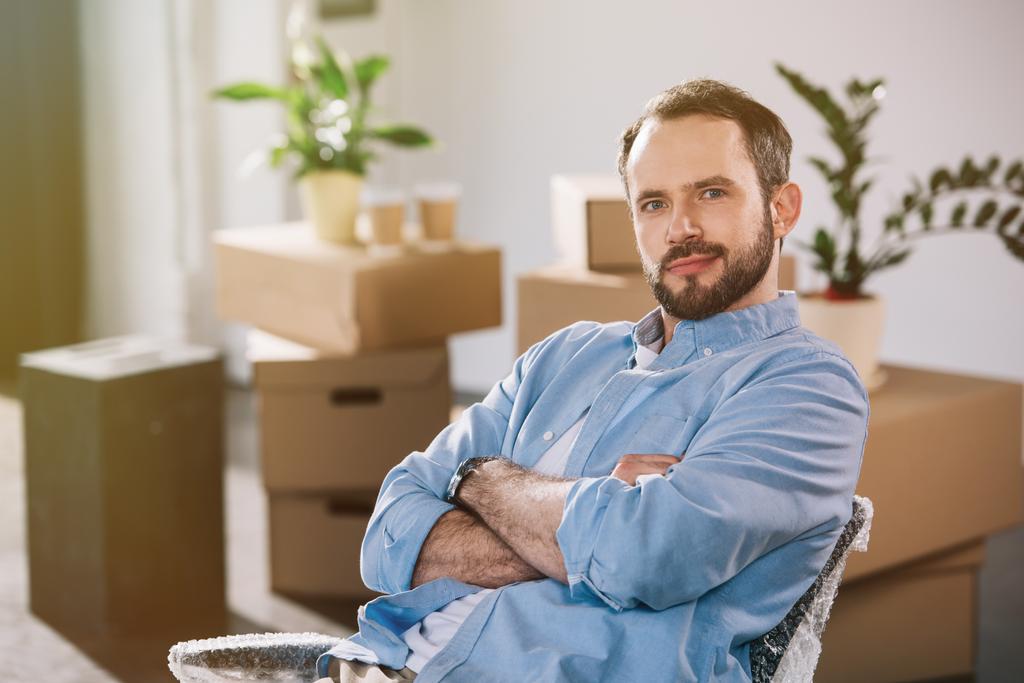handsome bearded man sitting with crossed arms and looking at camera while relocating in new office - Photo, Image