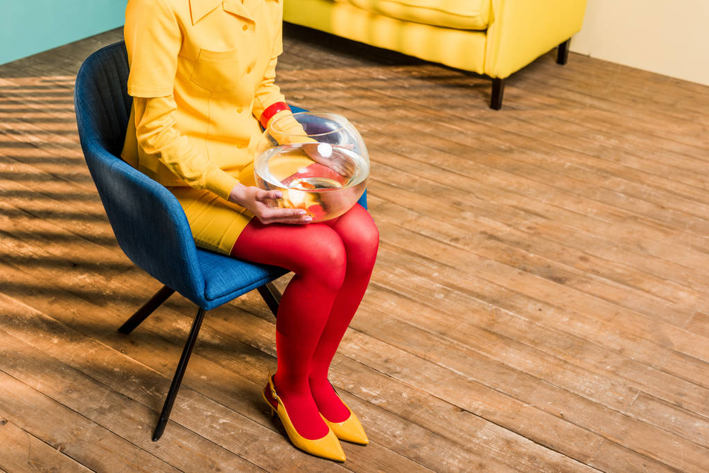 partial view of woman in retro clothing with golden fish in aquarium sitting on chair at colorful apartment, doll house concept - Photo, Image