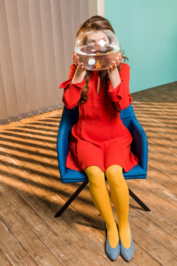 vista oscura de la mujer con peces de oro en el acuario sentado en la silla en el apartamento de colores, concepto de casa de muñecas
 - Foto, imagen