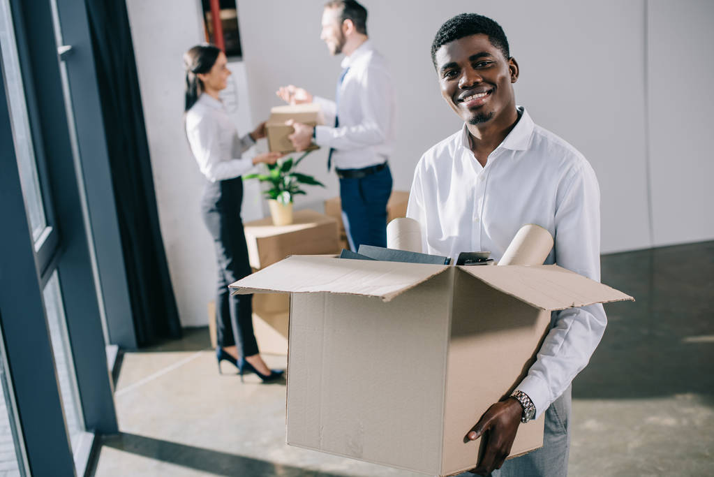 hombre de negocios afroamericano sosteniendo la caja de cartón y sonriendo a la cámara mientras sus colegas se quedan atrás en una nueva oficina
 - Foto, imagen