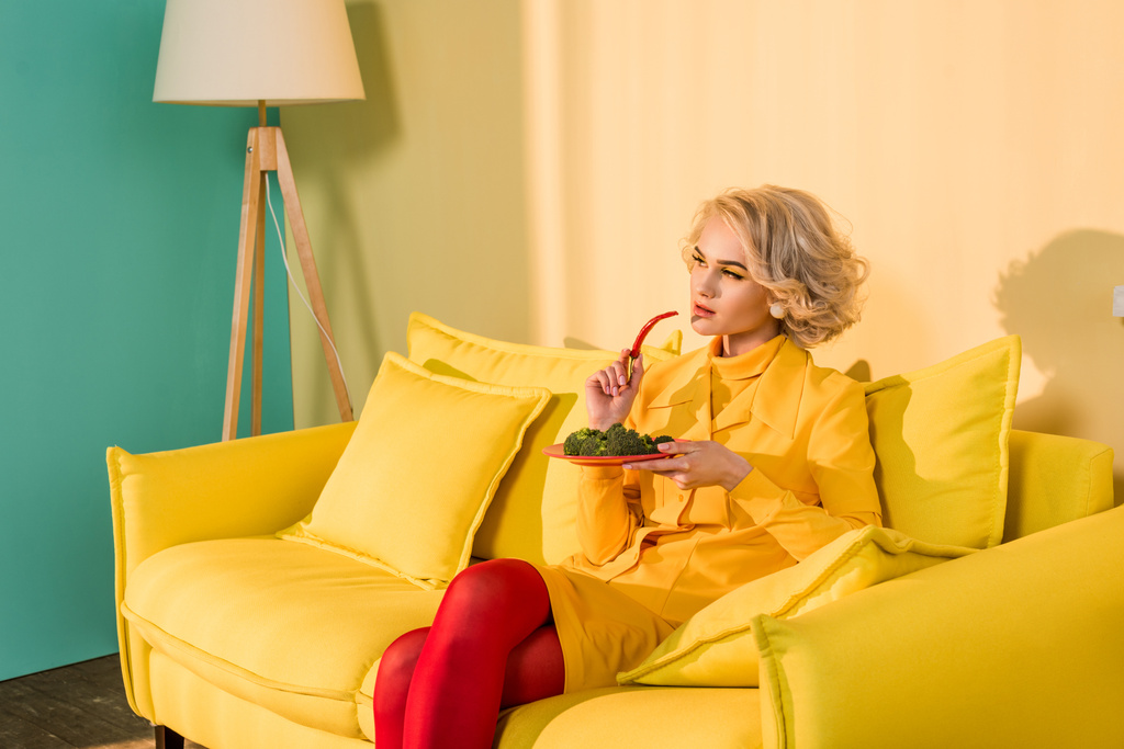 ρετρό στυλ γυναίκα με μπρόκολο σε πιάτο και τσίλι πιπέρι στο χέρι ακουμπά στον καναπέ στο φωτεινό διαμέρισμα, κούκλα σπίτι έννοια - Φωτογραφία, εικόνα