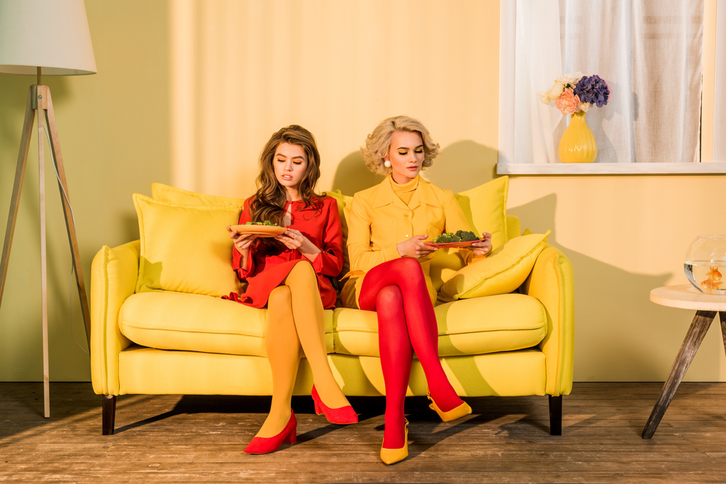 jolies femmes dans des vêtements rétro avec des légumes sur des assiettes assises sur un canapé jaune dans une pièce lumineuse, concept de maison de poupée
 - Photo, image