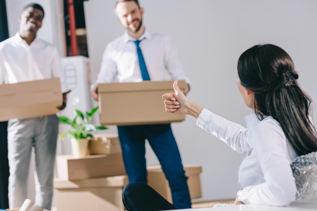 деловая женщина показывает большой палец коллегам мужского пола, держащим картонные коробки в новом офисе
 - Фото, изображение