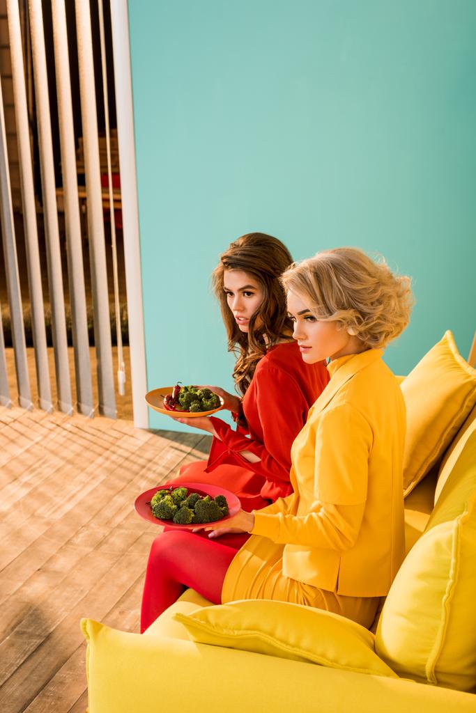 jolies femmes dans des vêtements rétro avec des légumes sur des assiettes assises sur un canapé jaune dans une pièce colorée, concept de maison de poupée
 - Photo, image