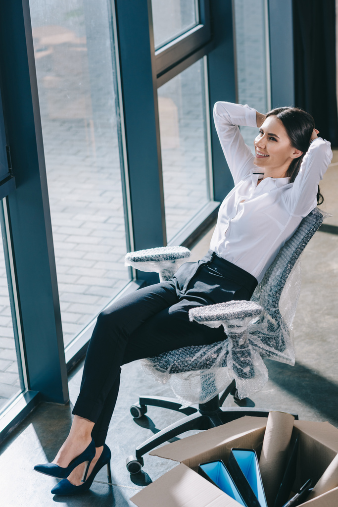Blick aus der Vogelperspektive auf eine glückliche junge Geschäftsfrau, die mit den Händen hinter dem Kopf in ihrem neuen Büro sitzt und wegschaut - Foto, Bild