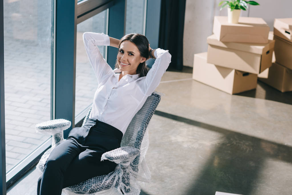 vista de ángulo alto de la joven mujer de negocios feliz sentado con las manos detrás de la cabeza y sonriendo a la cámara en la nueva oficina
 - Foto, Imagen