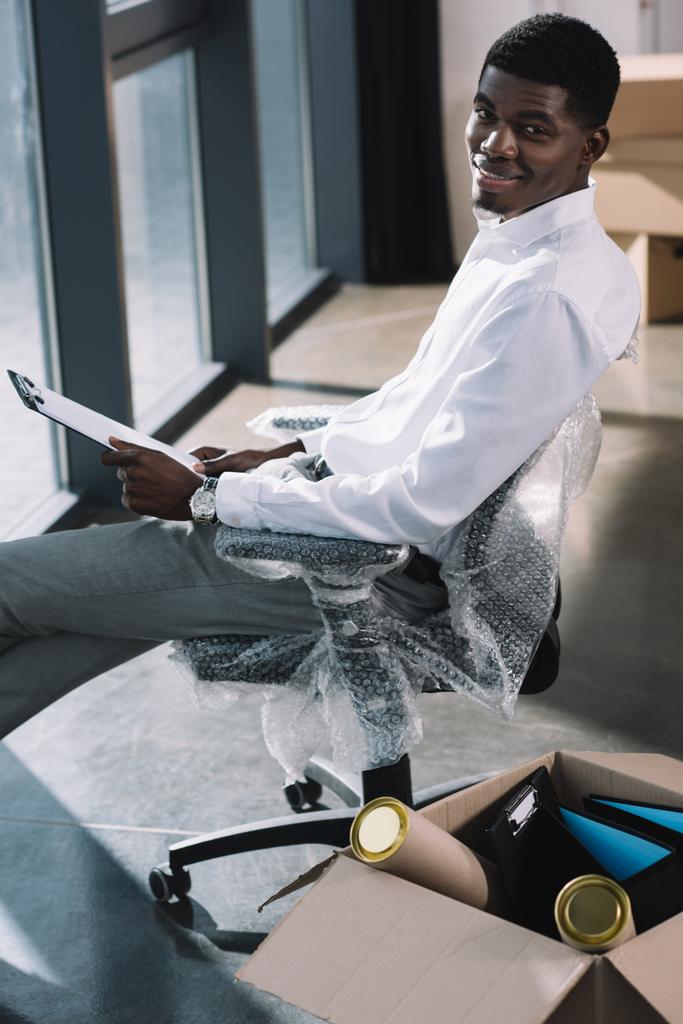 high angle view of african american businessman holding clipboard and smiling at camera while sitting in new office during relocation - Photo, Image