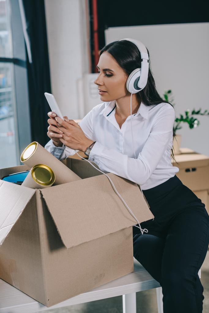 hermosa joven mujer de negocios en auriculares usando teléfono inteligente mientras desempaqueta la caja en una nueva oficina
   - Foto, Imagen