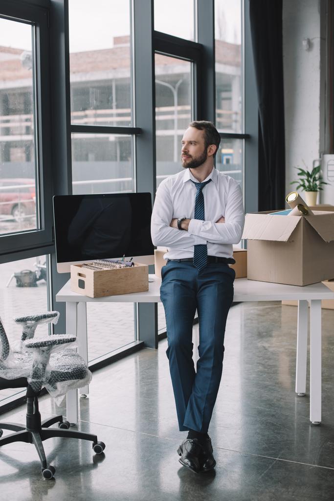 бизнесмен со скрещенными руками сидит на столе и смотрит вдаль в новом офисе
 - Фото, изображение