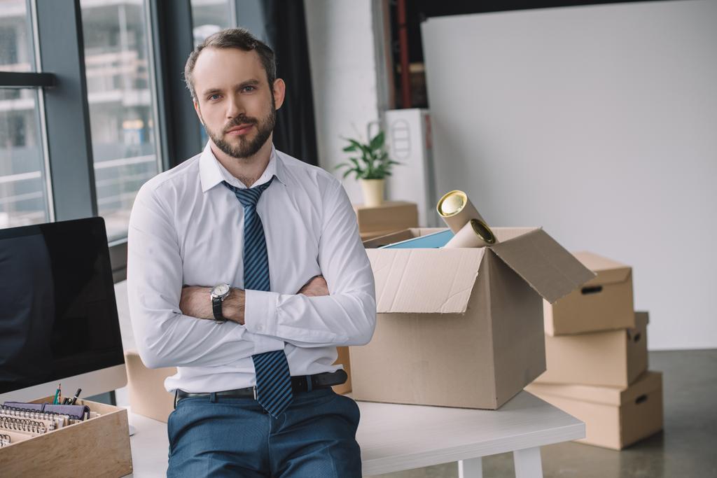 бизнесмен со скрещенными руками смотрит в камеру, сидя на столе с коробками в новом офисе
 - Фото, изображение