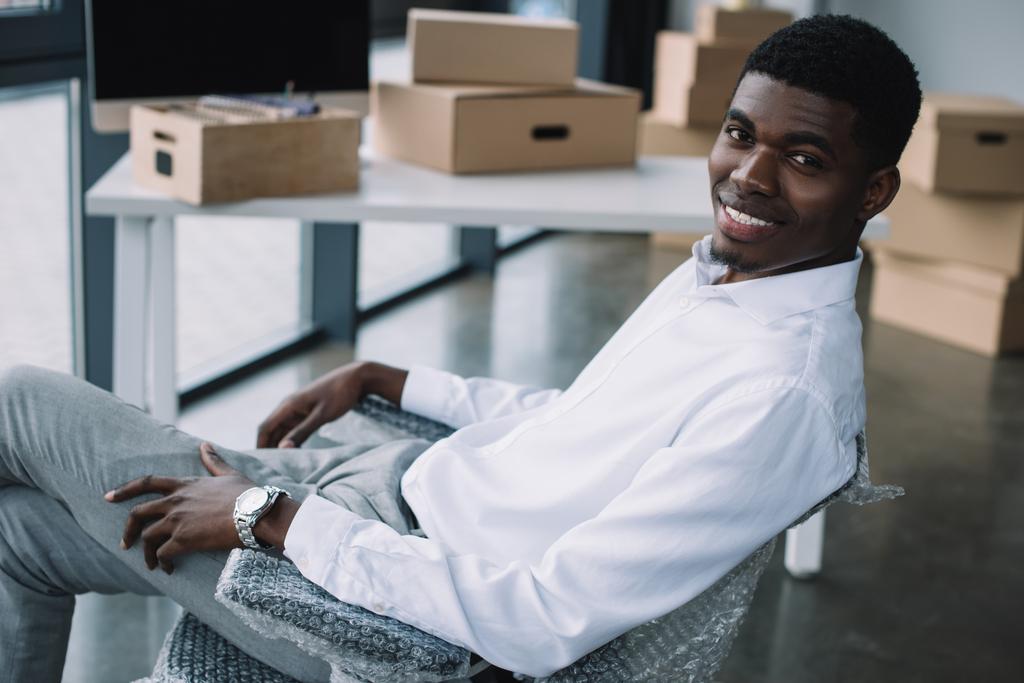 szczęśliwy młody biznesmen african american uśmiecha się do kamery siedząc w nowym biurze podczas relokacji - Zdjęcie, obraz