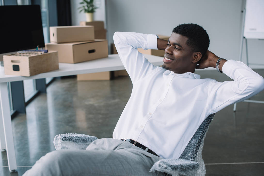 smiling young african american businessman sitting with hands behind head and looking away in new office - Photo, Image