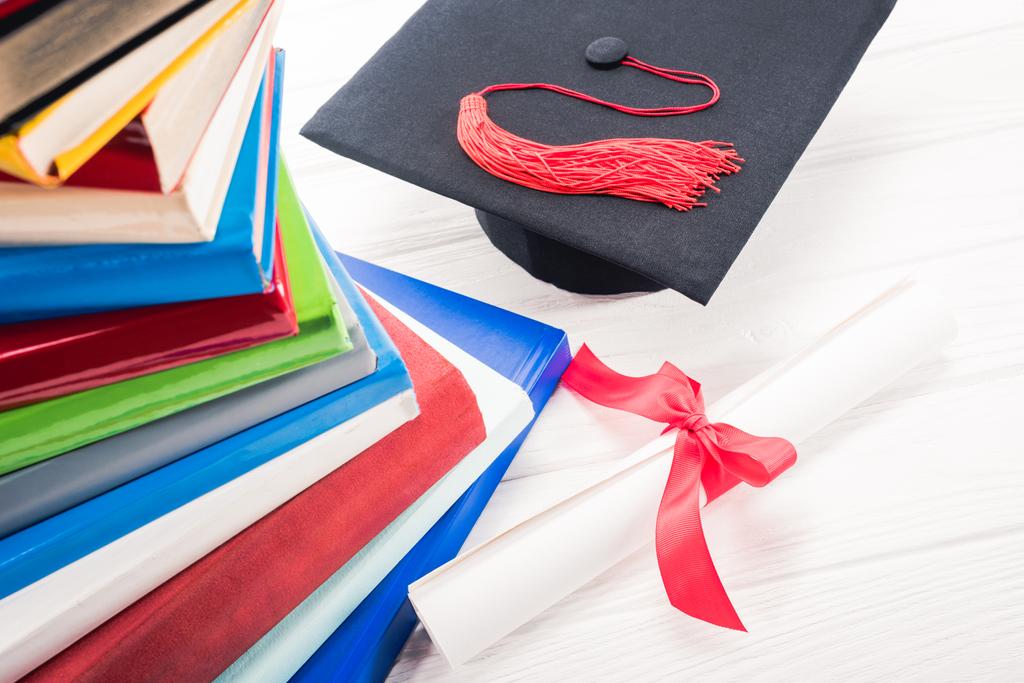 Chapéu de graduação e diploma na mesa branca por livros empilhados
 - Foto, Imagem