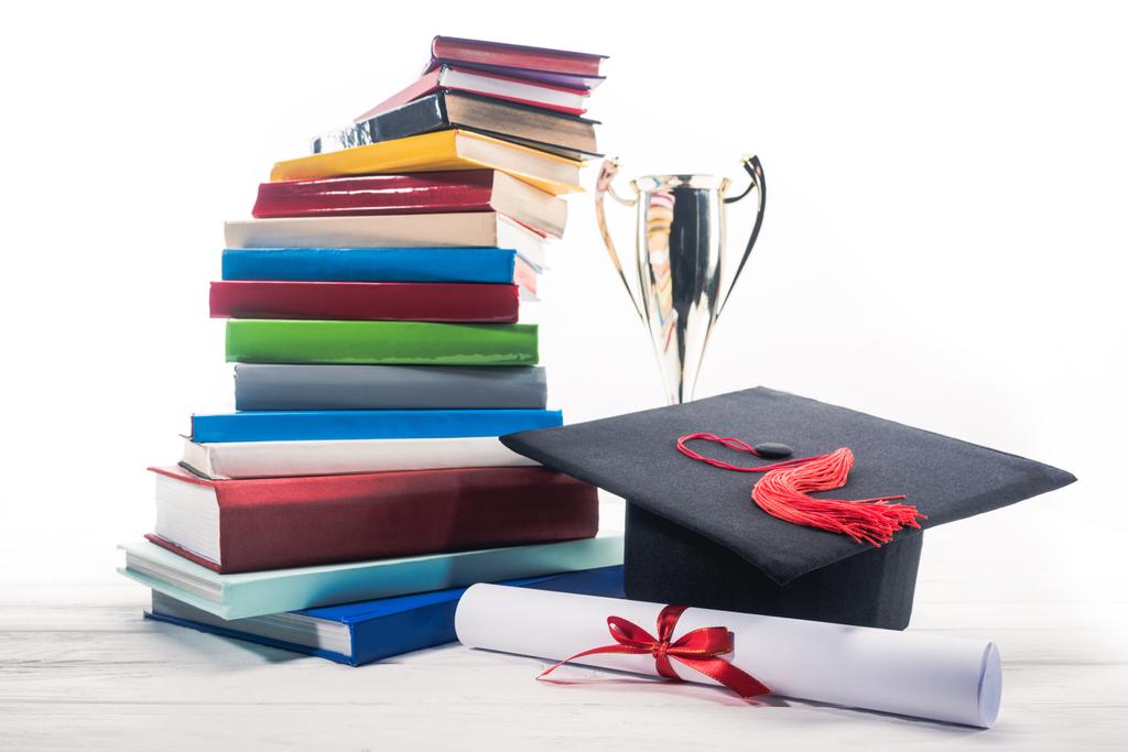 Graduation hat with diploma and trophy cup by stack of books - Photo, Image