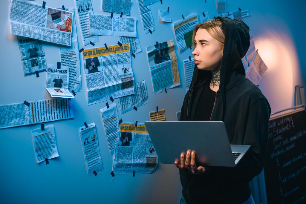 hacker mujer con portátil de pie frente a la pared con recortes de periódico
 - Foto, imagen