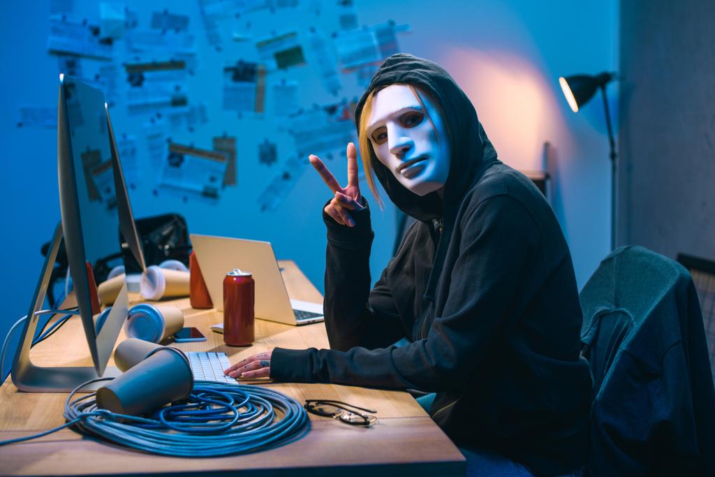 female hacker in mask showing v sign at workplace - Photo, Image